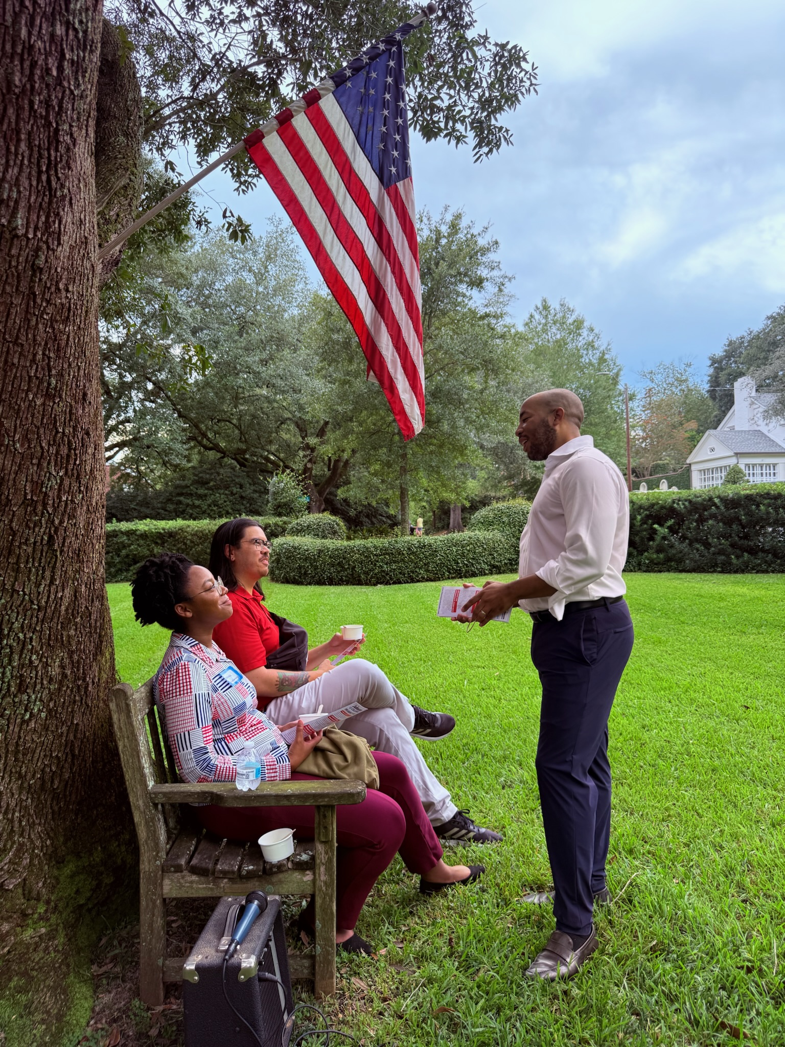 A group of three people engaged in a conversation outdoors under an American flag near a tree, with green grass in the foreground.