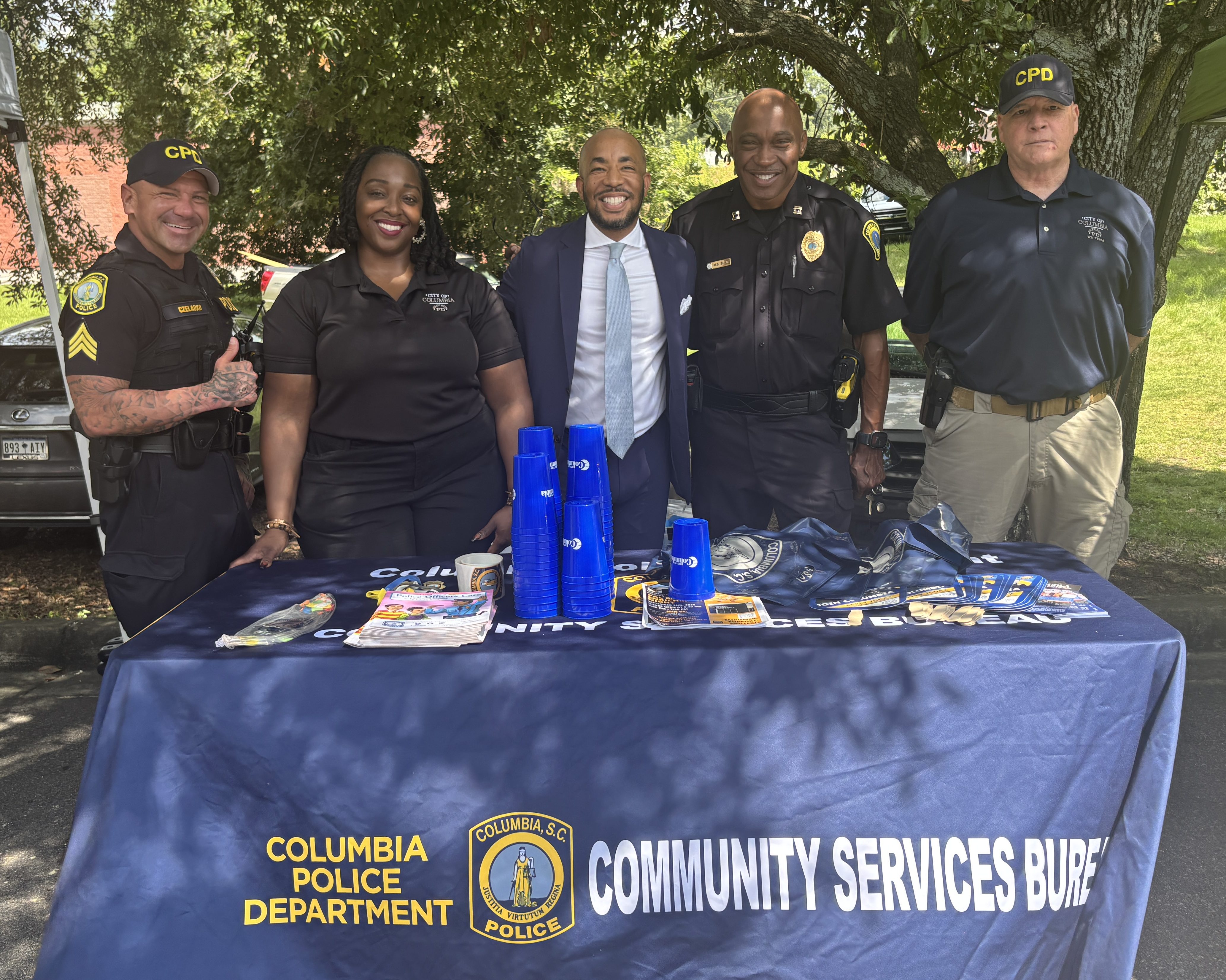 A group of police officers and a community member stand behind a table displaying materials for the Columbia Police Department's Community Services Bureau, outdoors under a tree.